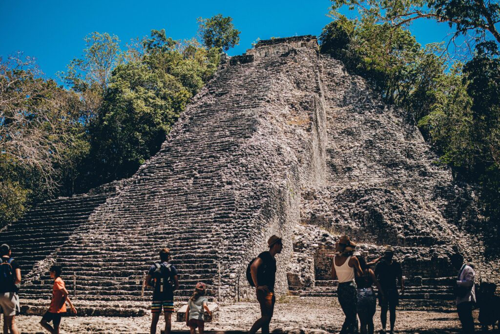 pexels photo 31456577 31456577 Visitors at the base of an ancient Mayan pyramid under a clear blue sky, surrounded by lush greenery.