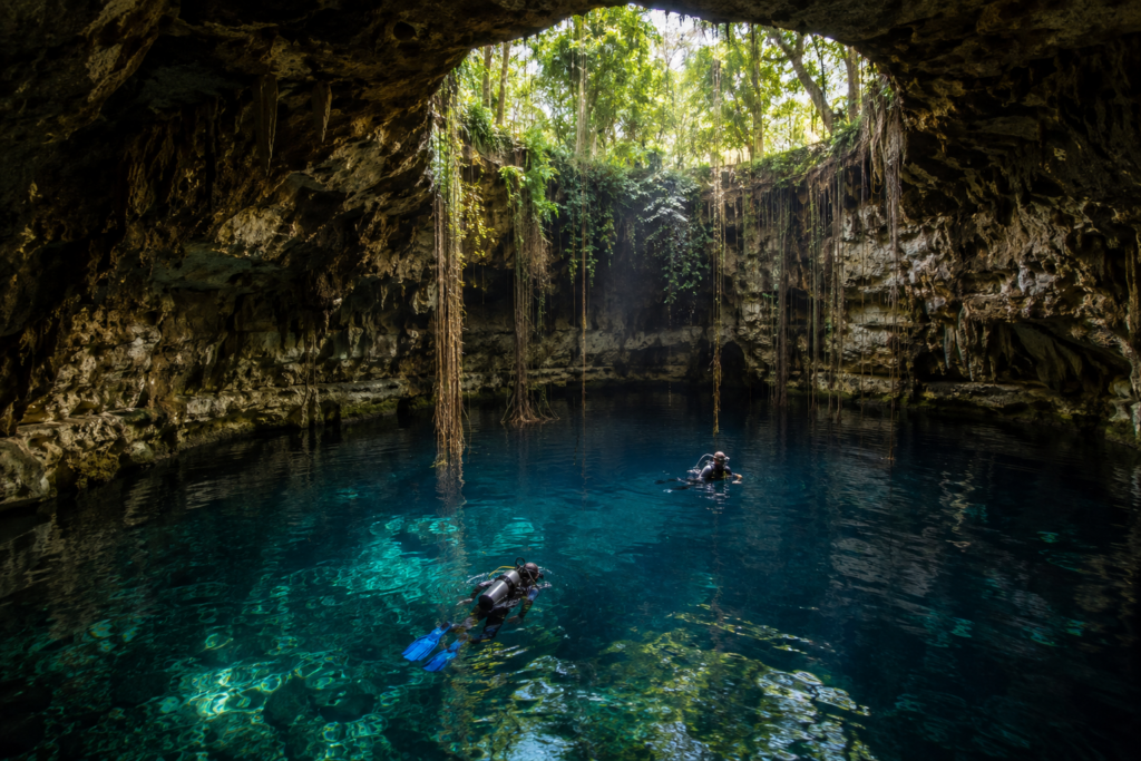 Plongée dans un cénote de la Riviera Maya Plongeurs explorant un cénote de la Riviera Maya dans une eau cristalline sous des rayons de lumière naturelle.
